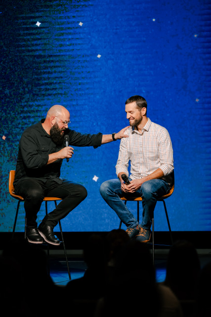 Man using a microphone praying for other main while sitting on stage