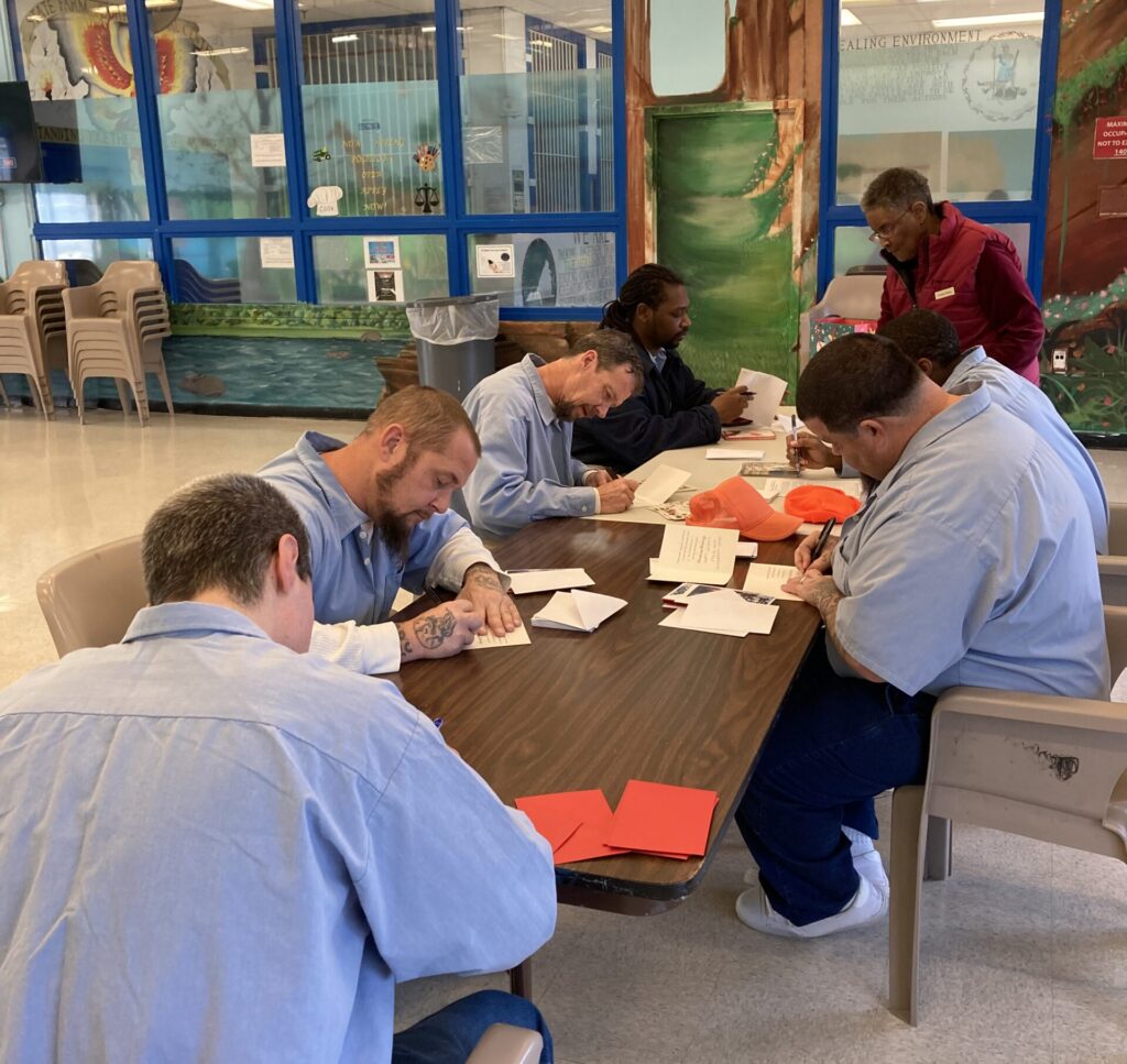 Chaplain supervises as inmates write cards at a table.