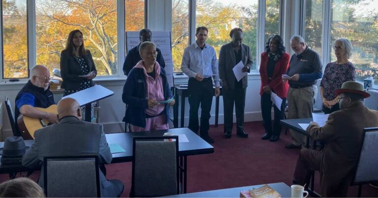 Chaplains singing together accompanied by a guitar in a room with large windows.